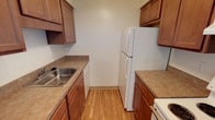 This single-floor residential unit offers a functional and straightforward layout designed for comfortable everyday living. The galley-style kitchen is efficiently arranged with warm medium-toned wood cabinetry lining both sides, providing ample upper and lower storage. Laminate wood flooring runs underfoot, complemented by neutral laminate countertops, a stainless steel sink, a white refrigerator, dishwasher, and range. The living area is generously proportioned, featuring soft carpet flooring, a textured ceiling, a ceiling fan, and a large sliding glass door dressed with vertical blinds that allows natural light to fill the space. A baseboard heating unit runs along the exterior wall. The bedrooms continue the carpeted flooring and feature textured ceilings, large sliding-door closets offering notable storage capacity, and neutral white walls that provide a clean, versatile backdrop. The bathroom is bright and clean, finished with classic white tile surrounding the tub-shower combination, a white vanity, and small mosaic tile flooring. Throughout the unit, the design is practical and understated, with consistent neutral tones and durable finishes that create a cohesive, livable environment.