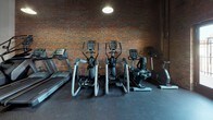 Located at 1650 Overbrook Rd in Richmond, VA, this single-floor fitness facility combines industrial character with functional workout space. The interior is defined by striking exposed brick walls, original heavy timber support columns, and soaring ceilings that lend the space an open, warehouse-inspired atmosphere. Pendant lighting fixtures hang throughout, casting warm, focused light across the rubber-floored workout areas.

The main floor is divided into distinct exercise zones. A dedicated cardio area lines one wall, housing a treadmill, multiple elliptical trainers, a stair climber, and a stationary bike — all set against the rich texture of the exposed brick backdrop. Full-length mirrors along one wall enhance the sense of space and provide visual feedback for workouts.

A separate strength training zone features a multi-station cable machine system, adjustable benches, and additional free weight equipment, offering a well-rounded resistance training environment. The rubber flooring throughout provides both safety and durability underfoot.

Natural light filters in through windows set into the brick walls, complementing the overhead pendant lighting. A private door leads to an adjoining corridor, and the space also includes a utility area with accessible storage. The overall ambiance is rugged yet purposeful, blending the historic character of the building's architecture with a fully equipped, modern fitness environment.