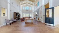 This single-story community clubhouse in Groton, Connecticut, offers a thoughtfully designed gathering space with a diverse range of amenities. The centerpiece is a grand great room boasting soaring vaulted ceilings with exposed beams, large arched windows that flood the space with natural light, rich wood-look tile flooring, and a striking stone fireplace. A decorative globe chandelier and vibrant artwork add character, while a generous leather sectional sofa and built-in display shelving create a warm, inviting atmosphere. Adjacent to the great room is a fully equipped kitchen featuring white cabinetry, granite countertops, a stainless steel refrigerator, mosaic tile backsplash, and a breakfast bar with seating — ideal for community gatherings. A dedicated conference room with a large table provides a quiet setting for meetings. The business center offers computer workstations and printing capabilities for residents' convenience. The fitness center is bright and spacious, lined with large windows overlooking the outdoor pool area, and is outfitted with treadmills, strength-training machines, and ceiling fans. A welcoming entryway with double glass doors and seating completes this well-rounded community facility.