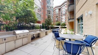Located at 1031 N Vermont St in Arlington, Virginia, this generously sized outdoor amenity terrace offers a well-appointed communal space designed for relaxation and entertaining. The terrace is finished with large-format porcelain tile pavers throughout, providing a clean and durable surface across its expansive footprint.

At the heart of the space is a built-in outdoor kitchen area, featuring multiple gas grills, a sink, and stone-finished countertops — ideal for group gatherings and al fresco cooking. High-top bar tables with blue upholstered seating are positioned nearby, offering a casual dining setting. A separate open-air dining zone with standard-height tables and chairs provides additional seating options for larger groups.

A lounge area with cushioned outdoor armchairs surrounds a fire pit, creating an inviting spot for relaxed conversation. Raised masonry planters filled with ornamental grasses and seasonal plantings add a natural, softening element to the hardscape and define distinct zones within the terrace.

The space is enclosed by wrought iron fencing and low masonry walls, framing views of the surrounding multi-story brick and glass residential buildings. Mature trees lining the perimeter provide shade and a sense of privacy. The overall ambiance is urban yet comfortable, making this terrace a functional and attractive outdoor amenity in the heart of Arlington.
