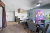 Located at 3402 Cahuenga Blvd W in Los Angeles, this single-floor amenity space is part of a contemporary multi-story residential complex. Accessed through full-length glass entry doors, the space opens into a welcoming courtyard surrounded by lush, mature planters featuring agave, bamboo, and flowering plants, lending a vibrant, garden-like atmosphere to the exterior approach.

Inside, the living room lounge area features soft blue-gray walls, patterned upholstered seating, carpeted floors, and colorful artwork, creating a comfortable and inviting gathering environment. A dedicated screening room adjoins the lounge, designed with tiered seating, plush cushioned sofas, a large flat-screen television, and warm earth-toned walls — ideal for communal viewing experiences.

The kitchen area is well-appointed with crisp white shaker cabinetry, light wood-look flooring, stone-finish countertops, stainless steel appliances including a full-size refrigerator, microwave, and sink, and teal accent walls that add a modern, polished touch. A pass-through counter connects the kitchen to the adjacent lounge, facilitating easy interaction between spaces.

The overall design balances contemporary finishes with warm, livable details, making this a functional and visually cohesive community space suited for socializing, relaxing, and entertainment.