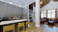 This single-floor communal space combines contemporary design elements with functional layout to create an inviting, multi-purpose environment. The kitchen bar area serves as a central focal point, featuring a sleek white countertop island paired with vibrant yellow bar stools that add a bold pop of color. Dark cabinetry lines the back wall, which is dramatically clad in a textured stacked-stone accent wall, adding depth and an industrial-chic character to the space. Pendant lighting hangs overhead, providing focused illumination over the bar area.

The flooring throughout is a warm wood-look material that ties the various zones together seamlessly. Structural white columns help define different areas while maintaining an open, flowing feel. Moving toward the lounge area, natural light pours in through large windows, illuminating a comfortable seating arrangement complete with patterned upholstery and accent rugs. A rich, dark wood-paneled ceiling detail in the lounge area adds warmth and contrast. The overall ambiance is modern yet welcoming, blending bold textures, warm tones, and contemporary furnishings into a cohesive and well-appointed communal setting.