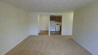 This single-floor space features a practical, open-concept layout that connects the main living area to the kitchen. The living area is finished with neutral-toned wall-to-wall carpeting, creating a clean and understated look, while the ceiling features a textured finish that adds subtle depth. The space is bright, with light-colored walls throughout that enhance the sense of openness.

The kitchen is accessible directly from the living area through a wide opening, allowing for easy flow between the two spaces. It is equipped with white appliances, including a range and oven, and features wood-toned upper cabinetry that provides ample storage. The kitchen floor transitions to a hard surface, offering a practical and easy-to-maintain finish distinct from the carpeted living area.

A hallway visible from the main living space leads further into the unit, suggesting additional rooms beyond what is immediately visible. A closet with bi-fold doors is also accessible from this hallway, providing convenient storage. Overall, the space presents a clean, functional environment with a simple, neutral palette that offers a versatile backdrop for a variety of furnishing styles.
