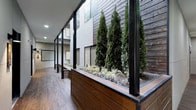 This single-floor space makes a strong architectural impression through its thoughtful blend of natural and industrial materials. A standout feature is the prominent built-in planter integrated into the interior corridor, housing tall, columnar evergreen trees and ornamental grasses that bring lush, living greenery into the built environment. The planter is clad in rich, dark wood paneling that complements the warm wood-tone flooring running throughout the space. Slender black steel columns and floor-to-ceiling glass partitions frame the corridor, creating a clean, modern aesthetic while allowing light to pass through and maintaining visual connectivity across different areas. The exterior facade, visible through the glazing, continues the dark wood cladding theme, reinforcing a cohesive material palette. Recessed ceiling lighting provides even, ambient illumination throughout the corridor. The overall design language is contemporary and sophisticated, combining the warmth of natural wood textures with the precision of steel and glass to create an environment that feels both refined and welcoming. The integration of live planting elements adds a biophilic quality that distinguishes this space and contributes to a calm, curated ambiance.