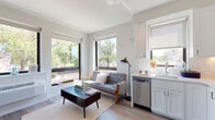 This thoughtfully designed single-floor unit offers a seamless blend of modern style and functional living. The open-concept living and kitchen area is bathed in natural light, thanks to multiple large black-framed windows that frame lush outdoor tree views and create an airy, welcoming atmosphere. Light-toned hardwood flooring runs throughout, tying the spaces together cohesively.

The kitchen is outfitted with crisp white shaker cabinetry, white subway tile backsplash, light countertops, and a full suite of stainless steel appliances including a gas range, over-the-range microwave, dishwasher, and refrigerator. A single-basin sink with a window above it adds both practicality and charm. The adjacent living area provides a comfortable retreat, with ample room for seating and a coffee table, all oriented to take advantage of the abundant natural light and street-level tree views.

The bedroom is a calm and restful space, featuring warm-toned walls, a ceiling fan, and generous proportions that allow for a full bedroom suite. Soft lighting from flanking table lamps adds to the relaxing ambiance.

The bathroom maintains the unit's clean aesthetic with a white vanity, undermount sink, chrome fixtures, a large mirror, and understated wall art, delivering a spa-like feel. Overall, this unit offers a polished, contemporary living experience with an emphasis on light, efficiency, and modern design.