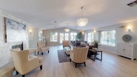 This single-floor space offers an open and airy layout designed for both comfort and style. The main living area features vaulted ceilings that enhance the sense of spaciousness, complemented by light hardwood flooring that runs throughout and contributes to the room's warm, welcoming ambiance. A statement capiz shell pendant chandelier anchors the space from above, while coordinating wall sconces provide soft, layered lighting throughout.

The living area is thoughtfully arranged with multiple seating zones, including a set of cream upholstered accent chairs with nailhead trim and a plush sectional sofa, offering versatile spaces for gathering or relaxing. A neutral color palette of soft grays and warm whites ties the furnishings together cohesively.

Natural light pours in abundantly through multiple large windows and a set of French doors, offering views of the surrounding greenery outside. The windows are dressed with clean white blinds, allowing flexible light control while maintaining the bright, open feel of the interior.

Overall, the space exudes a refined yet approachable aesthetic, balancing elegant design details with a comfortable, livable atmosphere suitable for a variety of uses.