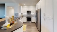 This well-appointed single-floor residence offers a thoughtfully designed open-concept layout that seamlessly connects the main living areas. The living room serves as the heart of the home, featuring rich hardwood-style flooring, a warm taupe accent wall, and floor-to-ceiling sliding glass doors that open onto a private balcony, flooding the space with natural light and providing views of the surrounding community.

The kitchen is a standout feature, outfitted with crisp white shaker-style cabinetry, dark gray countertops, and a striking stone-tile mosaic backsplash. Stainless steel appliances, including a gas range, built-in microwave, and French door refrigerator, round out this highly functional cooking space. A breakfast bar adds additional prep and dining space.

The bedrooms offer comfortable retreats; the primary bedroom includes a walk-in closet and a bold striped accent wall, while a secondary bedroom features neutral tones and a bright window with decorative curtains. The bathroom is finished with dark wood-style flooring, a dark-toned accent wall, a single vanity with dark countertop, and a shower-tub combination. The cohesive use of gold, taupe, and white tones throughout creates a warm, modern ambiance.