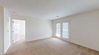 This single-floor residence offers a practical and well-organized layout designed to maximize functionality throughout. The main living area is generously sized, featuring neutral beige carpeting, crisp white walls, and a pair of French doors fitted with blinds that allow for natural light while providing outdoor access. An open passage connects the living space to a galley-style kitchen, which is outfitted with warm wood cabinetry, light countertops, a double stainless steel sink, and a white electric range. A dedicated laundry area is accessible directly from the kitchen and accommodates a full-size washer and dryer side by side. Adjacent to the main living area, a hallway leads to the private rooms. The bedroom features the same neutral carpeting and direct access to a generously proportioned walk-in closet equipped with wire shelving and hanging rods along two walls, offering ample storage capacity. The bathroom is finished with tile flooring and includes a full-size soaking tub with an overhead shower, a toilet, and ventilation. Throughout the unit, white walls and consistent flooring materials create a cohesive, clean aesthetic.