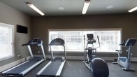 This single-floor fitness room is designed to support cardiovascular exercise in a functional, no-frills environment. The space is equipped with two treadmills and an elliptical trainer, positioned along a wide, horizontal window that spans much of one wall. The window, fitted with horizontal blinds, allows ample natural light to filter into the room, creating a bright and motivating workout environment while offering a view of the outdoors.

The walls are finished in a deep charcoal-gray tone, lending a modern, focused atmosphere to the space. Overhead, fluorescent strip lighting supplements the natural light, ensuring the room remains well-lit throughout the day. The flooring consists of durable, dark-toned material suited for fitness use, providing stability and easy maintenance underfoot.

The ceiling is fitted with a recessed speaker and a ceiling fan, contributing to both comfort and air circulation during workouts. The layout is efficiently organized, allowing users to move comfortably between machines. Overall, the space offers a clean, practical setting for cardiovascular training.