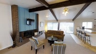 This single-floor space makes a strong first impression with its distinctive architectural character. The living area is anchored by dramatic exposed dark wood ceiling beams that add warmth and a rustic charm to the open layout. A ceiling fan with a light fixture provides both comfort and illumination, complementing the abundant natural light that streams in through large sliding glass doors, which also offer a pleasant view of the outdoors.

The room's focal point is a full-height brick fireplace set against a bold blue accent wall, creating a striking visual contrast. A wall-mounted flat-screen television sits above the fireplace surround, seamlessly blending entertainment with the room's classic aesthetic. The carpeted living area is comfortably furnished with a plush sofa and a pair of striped accent chairs arranged around a round coffee table, creating an inviting and functional seating arrangement.

The overall ambiance of the space is one of relaxed comfort with character — the combination of exposed beams, brick masonry, and soft neutral carpeting gives the interior a cozy yet spacious feel, while the generous glazing ensures the space remains bright and connected to the outdoors throughout the day.