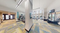 Located at 805 North Olive Avenue in West Palm Beach, Florida, this fitness center offers a spacious and thoughtfully designed workout facility spread across a single floor. The space is divided into two distinct zones that together provide a comprehensive exercise environment.

The cardio area is immediately striking, featuring soaring double-height ceilings with rows of large clerestory windows that flood the room with natural light and offer open views of the surrounding landscape. Multiple treadmills, ellipticals, and stationary bikes are arranged along the windowed walls, while full-length mirrors on one side visually expand the space further. Pendant and linear LED lighting supplements the abundant natural light for a bright, energizing atmosphere.

The strength and functional training zone is anchored by a bold teal curved accent wall, warm wood-toned columns, and wall-mounted flat-screen televisions. This area is outfitted with cable machines, a Life Fitness functional trainer, TRX suspension equipment, stability balls, free weights, and a variety of resistance machines. Glass-panel sliding doors connect the zones, maintaining visual openness between areas.

Throughout the facility, a distinctive herringbone-patterned carpet in blue, yellow, and gray tones adds a modern, contemporary aesthetic that ties the two areas together cohesively.
