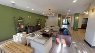 This single-floor space presents a stylish and inviting communal lounge area that blends modern design with comfortable functionality. The room is anchored by bold olive-green accent walls that create a rich, warm contrast against the light neutral tones of the ceiling and flooring. Wide-plank light wood flooring runs throughout, lending a clean and cohesive aesthetic to the space.

A striking multi-arm brass chandelier serves as a focal point overhead, casting warm, ambient light across the seating area below. The lounge is furnished with a curated mix of cream upholstered sofas and armchairs, a rich brown leather sofa accented with navy throw pillows, and a dark wood coffee table — together creating a layered, sophisticated sitting area arranged around a patterned area rug.

Along the accent wall, an industrial-style metal shelving unit and a dark wood sideboard provide both decorative and functional storage, displaying books, plants, and decorative objects. Recessed ceiling lighting supplements the chandelier, ensuring the space is well-lit throughout.

A long, naturally lit corridor extends toward the back of the space, where glass-paneled doors allow views to an outdoor area, enhancing the sense of openness and connecting the interior to the exterior environment. The overall design balances elegance with approachability, making this a welcoming space for socializing or quiet relaxation.