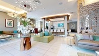 This thoughtfully designed single-floor amenity space offers a seamless blend of social, recreational, and functional zones unified by a cohesive contemporary aesthetic. The main lounge area anchors the space with generously sized taupe leather sofas accented by teal throw pillows, arranged around a white lacquer coffee table atop a soft area rug. Floor-to-ceiling windows flood this seating area with natural light while offering views of the surrounding outdoor greenery. A striking decorative screen partition with an organic, cutout pattern serves as a visual divider between lounge zones.

The central bar area is a standout feature, defined by a curved, wood-paneled ceiling with warm integrated uplighting. It includes a granite countertop, warm wood cabinetry, a beverage refrigerator, a wall-mounted TV, and shimmering mosaic tile backsplash. White bar stools line the counter, creating an inviting social hub.

Adjacent to the bar, a dedicated workstation area provides computer terminals for practical use. The game room features a full-size billiards table and a shuffleboard table beneath a cluster of modern pendant lights, with patterned tile flooring adding visual texture. Throughout the space, recessed lighting and designer fixtures contribute to a polished, resort-style ambiance.