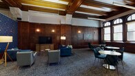 This distinguished single-floor space masterfully combines classic architectural heritage with contemporary design. The main lounge area is defined by soaring, exposed wood-beamed ceilings with gilded accents, rich dark wood wall paneling, and wall-mounted sconces that cast a warm, refined glow throughout. A row of arched windows along one wall floods the space with natural light while offering views of the surrounding cityscape.

The layout is thoughtfully divided into distinct zones. A lounge area features plush blue velvet sofas and modern gray upholstered chairs arranged around low tables, alongside a wall-mounted flat-screen television. A separate seating zone showcases bold navy geometric-patterned wallcovering paired with compact velvet chairs and additional televisions. Multiple high-top and standard café-style tables with dark upholstered seating provide flexible options for casual gatherings or meetings.

A well-appointed kitchenette serves the space, featuring navy cabinetry with brushed gold hardware, white quartz countertops, a undermount sink with a champagne bronze faucet, stainless steel appliances, and stone tile flooring. A quieter secondary lounge area with neutral upholstered furniture and its own television offers a more intimate retreat. Patterned carpeting runs throughout, tying the varied zones together cohesively.