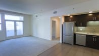 This single-floor space showcases a thoughtfully designed open-concept layout that integrates the living area and kitchen into one cohesive environment. The living area is finished with soft, neutral-toned carpeting that adds warmth underfoot, while the kitchen zone transitions to tile flooring, clearly delineating the two functional spaces. Large windows along the living area allow natural light to fill the room, enhancing the bright, airy atmosphere created by the crisp white walls and smooth ceilings fitted with ventilation registers.

The kitchen is appointed with dark espresso cabinetry that contrasts elegantly against the lighter surroundings. A full-size stainless steel refrigerator anchors the kitchen, complementing the contemporary aesthetic. A countertop workspace is partially visible, suggesting practical functionality within a compact footprint.

A hallway visible through an open doorway leads further into the unit, indicating additional rooms beyond the main living space. The overall design emphasizes clean lines, modern materials, and a neutral color palette, creating a versatile and move-in-ready environment suited to a variety of interior styles.