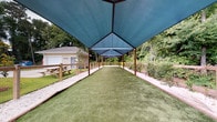 Located at 3112 Maybank Hwy on Johns Island, SC, this single-story outdoor facility is thoughtfully designed for active use in a natural lowcountry setting. The centerpiece of the property is a generously sized covered turf area, sheltered by a large blue shade canopy supported by sturdy metal posts. The canopy runs the full length of the turf corridor, providing substantial protection from sun and light rain while maintaining an open-air feel on all sides. The artificial turf surface is well-maintained and bordered by sandy pathways, with wooden fencing and wire mesh enclosing the perimeter to define the space clearly.

Adjacent to the covered turf area is an open sandy yard, also enclosed by wooden post-and-wire fencing, offering additional outdoor space. A compact, single-story support building with light-colored lap siding and a shingled roof sits nearby, equipped with exterior utility connections. A storage container is also present on the grounds. Picnic tables are positioned near the building, providing gathering spots.

The entire property is surrounded by mature trees and natural vegetation, creating a secluded, wooded ambiance characteristic of the Johns Island landscape. The combination of shaded turf, open sand areas, and functional infrastructure makes this a versatile outdoor facility.