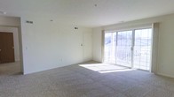 This single-floor space offers a clean, open layout designed for comfortable everyday living. The main living area is finished with wall-to-wall carpeting in a neutral tone, providing a warm and inviting foundation throughout the room. Crisp white walls and a smooth white ceiling enhance the sense of openness, while a ceiling-mounted smoke detector and HVAC vent reflect practical, well-maintained infrastructure.

A standout feature of the living area is the large sliding glass door, which floods the space with generous natural light and opens directly onto a private balcony with a white railing. The outdoor view beyond the balcony offers a glimpse of the surrounding neighborhood, adding a pleasant sense of connection to the outdoors.

An interior door provides access to additional areas of the unit, and a wall-mounted thermostat or control panel is conveniently positioned nearby. Baseboard elements along the lower walls contribute to the room's functional design. The overall ambiance is bright, airy, and move-in ready, with a neutral palette that offers a versatile backdrop for a variety of interior styles and furnishings.