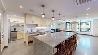 This single-floor space in Walnut Creek, California is thoughtfully designed to support both culinary and communal activities. The main kitchen serves as a central highlight, anchored by an expansive granite island with seating for multiple guests along one side. Cream-toned shaker cabinetry lines the perimeter walls, complemented by white vertical tile backsplash and under-cabinet lighting. Globe pendant lights with brass hardware hang above, adding a warm, contemporary touch. Polished concrete flooring flows throughout, lending an industrial-modern aesthetic.

A secondary galley-style prep kitchen offers additional workspace, featuring dark stone countertops, light maple cabinetry, open floating shelves, and full-size stainless steel refrigeration — ideal for high-volume food preparation.

The dining and gathering area accommodates a large wood communal table surrounded by woven chairs, set atop a vibrant, patterned area rug. A wall-mounted television, a sculptural circular mirror, and a distinctive geometric ceiling feature with integrated linear lighting contribute to the room's character. A projector is also available for presentations or screenings.

A glass-paneled entry door connects the interior to an adjacent outdoor patio, allowing natural light to filter in and providing a seamless transition between indoor and outdoor settings.