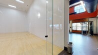This fitness center is a comprehensive, full-featured facility designed to accommodate a wide range of workout routines. The main gym floor is expansive and open, with soaring double-height ceilings accentuated by bold terracotta-red walls, rich wood paneling, and exposed industrial ductwork that lend the space a modern, energetic aesthetic. Large windows on multiple walls flood the interior with natural light while offering views of the surrounding landscape, and suspended linear lighting ensures consistent illumination throughout. The floor is finished with durable rubber sport flooring ideal for high-intensity training. Equipment is plentiful and well-organized, including free weight racks, barbells, benches, cable machines, weight stacks, kettlebells, medicine balls, and a variety of cardio machines. Full-length mirrors line sections of the walls, enhancing spatial depth and supporting form-focused training. A metal staircase with cable railings leads to an upper mezzanine level that provides additional equipment storage and workout space. Adjacent to the main floor, a glass-walled racquetball court features a pristine hardwood playing surface, bright white walls, and overhead fluorescent lighting, offering a dedicated space for court sports and high-intensity play.