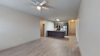 The Stables offers a well-designed single-story unit that combines functionality with a clean, contemporary aesthetic. The open-concept living and kitchen area forms the heart of the space, where light hardwood-style flooring extends seamlessly throughout, creating a sense of continuity and warmth. A ceiling fan with integrated lighting anchors the living area, providing both comfort and illumination.

The kitchen is positioned centrally, featuring a dark-toned island that serves as a natural dividing point between the living and cooking areas. Medium-toned cabinetry lines the kitchen walls, complemented by stainless steel appliances, including a full-size refrigerator, and a built-in microwave above the range. The layout promotes an efficient workflow while remaining visually open to the rest of the unit.

A hallway visible beyond the kitchen leads to additional rooms, suggesting a practical and well-organized floor plan. The entry door, finished in a classic paneled style, is conveniently accessible from the main living space. Neutral wall tones throughout contribute to a bright, versatile ambiance well-suited for a variety of interior styles.