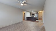 The Stables offers a well-designed single-story unit that combines functionality with a clean, contemporary aesthetic. The open-concept living and kitchen area forms the heart of the space, where light hardwood-style flooring extends seamlessly throughout, creating a sense of continuity and warmth. A ceiling fan with integrated lighting anchors the living area, providing both comfort and illumination.

The kitchen is positioned centrally, featuring a dark-toned island that serves as a natural dividing point between the living and cooking areas. Medium-toned cabinetry lines the kitchen walls, complemented by stainless steel appliances, including a full-size refrigerator, and a built-in microwave above the range. The layout promotes an efficient workflow while remaining visually open to the rest of the unit.

A hallway visible beyond the kitchen leads to additional rooms, suggesting a practical and well-organized floor plan. The entry door, finished in a classic paneled style, is conveniently accessible from the main living space. Neutral wall tones throughout contribute to a bright, versatile ambiance well-suited for a variety of interior styles.