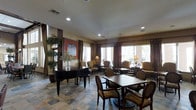 This single-floor communal space is thoughtfully designed to serve as both a social gathering area and a refined lounge. The room features a striking black grand piano positioned prominently near the entrance, lending the space a sophisticated, upscale character. Natural light pours in through multiple large windows dressed with rich brown curtain panels, illuminating the interior and offering views of the surrounding outdoor greenery and architecture.

The flooring consists of large-format stone tiles in warm, earthy tones, adding texture and a sense of permanence to the space. Recessed ceiling lighting supplements the natural light, ensuring the room remains well-lit throughout. Seating is arranged in distinct zones: cushioned armchairs are grouped near the windows to create intimate conversation areas, while dining-style tables surrounded by striped upholstered chairs occupy the central floor space, accommodating group gatherings.

Decorative elements include lush potted plants, table lamps with warm-toned shades, and a framed artwork piece displayed on the wall, contributing to the overall inviting ambiance. The combination of elegant furnishings, natural light, and thoughtful layout makes this a versatile and welcoming communal environment.