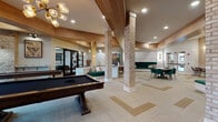 Located at 1399 Herrington Rd in Lawrenceville, GA, this single-story clubhouse offers a thoughtfully designed, open-concept layout that seamlessly blends social, recreational, and professional spaces. The interior is defined by a striking mix of warm wood ceiling beams, whitewashed brick columns, and light hardwood flooring accented by patterned carpeting in neutral tones — creating an inviting, upscale atmosphere throughout.

The main gathering area serves as the centerpiece of the clubhouse, featuring a round poker-style game table surrounded by rich emerald green velvet chairs, alongside built-in bench seating upholstered in the same bold green. Multiple seating arrangements accommodate both casual lounging and group gatherings. Pendant chandeliers and recessed lighting provide warm, layered illumination throughout.

The game room is well-appointed with a full-size billiards table and a shuffleboard table, complemented by exposed brick accent walls and framed game-themed artwork. Large windows flood the space with natural light and offer views of the outdoors.

A dedicated business center provides individual workstations with computers and a printer, set against wood-paneled walls adorned with large-format artwork. A marble-topped desk with stylish seating adds a refined touch to this functional area. Together, these spaces make the clubhouse a versatile and welcoming community hub.