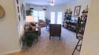 This inviting living space is thoughtfully designed to balance comfort and style across a single floor. The main living area is anchored by plush wall-to-wall carpeting that adds warmth underfoot, while large windows dressed with patterned curtains allow generous natural light to fill the room. A ceiling fan overhead provides both comfort and a subtle decorative touch. French doors lead to an outdoor area, further enhancing the sense of openness and connectivity to the outside. The living room is furnished with a comfortable neutral-toned sofa and armchair arrangement, complemented by a sleek dark coffee table and a coordinating dark accent chair. A leaning ladder shelf on one side of the room provides functional display space, showcasing decorative accents and greenery that bring a natural, lively energy to the interior. Potted plants are strategically placed throughout, adding a fresh, organic element to the overall aesthetic. The layout is open and spacious, allowing for easy movement and a welcoming flow between seating areas. Overall, the space exudes a warm, well-appointed ambiance that is both practical and visually appealing, making it an ideal environment for relaxing or entertaining.
