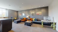 This single-floor space is thoughtfully designed as a contemporary lounge and business center, combining comfort and functionality in a cohesive, modern aesthetic. The lounge area is anchored by plush gray sofas and vibrant orange armchairs accented with teal and yellow throw pillows, creating a lively yet sophisticated seating arrangement. Lattice-style coffee tables in a dark finish add a geometric element, while a coordinating patterned carpet in muted gray with orange accents ties the space together. Recessed lighting illuminates the room evenly overhead, highlighting a gallery-style arrangement of bold, abstract artwork displayed on textured gray walls that runs the length of the lounge. A sleek desk with an iMac workstation provides a dedicated business area, positioned against a striking accent wall featuring textured wallpaper in a mosaic pattern with a bold olive-green vertical stripe. An artistic wall piece and a second monitor setup reinforce the space's dual-purpose nature. Natural light filters in through a sheer-curtained window near the workstation, adding warmth to the overall atmosphere. The space flows openly between the social lounge and the work area, making it well-suited for both collaborative gatherings and focused productivity.