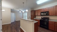 Located at 120 Cinema Dr in Hendersonville, Tennessee, this single-floor residence offers a thoughtfully designed open-concept layout that seamlessly connects the kitchen and living areas. The kitchen is a standout feature, showcasing rich medium-toned cabinetry, granite-look countertops, and a central island with a built-in sink and pendant lighting overhead. Black appliances, including a range, microwave, and refrigerator, complement the warm cabinetry tones. Dark hardwood-style flooring runs throughout the main living and kitchen areas, adding a sense of warmth and continuity. The living area benefits from natural light through blinds-fitted windows and is topped with a ceiling fan for added comfort. A carpeted room with its own exterior door and transom window offers versatile use as a bedroom or flex space, with a ceiling fan and ample natural light. The bathroom is equipped with a single-sink vanity featuring warm wood cabinetry, a full tub-shower combination with tiled surround, and a generous overhead storage cabinet. The overall design strikes a balance between functionality and comfort, with neutral wall tones and consistent finishes creating a cohesive, inviting atmosphere throughout the space.