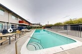 Located at 1611 Cormorant Way in Sacramento, California, this outdoor community pool area serves as a central amenity within a residential complex. The space is defined by a generously sized rectangular swimming pool with clear turquoise water, accessible via stainless steel entry rails and marked with depth indicators along its edges. The pool deck is finished in smooth concrete and provides ample room for recreation and relaxation. A dedicated lounge area is situated along one side of the pool, furnished with chaise lounge chairs and small tables, offering a comfortable setting for sunbathing and socializing. The entire pool area is enclosed by a sturdy metal perimeter fence, providing both safety and privacy. A gated entry point with a brick accent column adds a touch of character to the enclosure. The surrounding complex features a two-story building with a modern exterior accented by warm wood-toned balcony panels, navy blue trim, and staircase access between levels. Adjacent green lawn areas and mature trees add a refreshing natural element to the setting. A covered carport parking area is also visible along the perimeter. The overall ambiance is functional and inviting, offering residents a well-maintained outdoor retreat well-suited to Sacramento's warm climate.