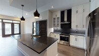 Located in Decatur, GA, this single-floor residence showcases a thoughtfully designed open-concept layout with contemporary finishes throughout. The kitchen is a standout centerpiece, featuring a large island with a dark quartz countertop, an integrated sink, and striking pendant lights overhead. White shaker-style cabinetry with black hardware lines the walls, complemented by a bold black penny-tile backsplash, dark countertops, and a full suite of stainless steel appliances including a range, range hood, and French door refrigerator. Upper glass-front cabinets and a built-in wine rack add refined detail. The kitchen flows seamlessly into the living area, which is bathed in natural light through large French doors that open to a private balcony. Hardwood-style flooring runs throughout the main living areas, lending warmth and continuity. The primary bathroom offers a clean, spa-like aesthetic with white subway tile, a glass-enclosed shower, a soaking tub, dark quartz vanity countertop, and direct access to a well-organized walk-in closet with built-in shelving. Additional bedrooms feature plush carpeting, large windows, and ceiling fans. The overall ambiance is modern and polished, with a cohesive contrast of black and white design elements.