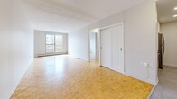 This well-organized 2-bedroom, 1-bathroom unit is laid out across a single floor, making efficient use of its space. The living area is a standout feature, showcasing warm parquet wood flooring in a classic herringbone-style pattern that extends throughout most of the unit. A large window at the end of the living room draws in natural light and frames views of surrounding trees and neighboring architecture, lending the space an airy, open feel. A baseboard heating system is present along the walls. The primary bedroom is generously sized, offering a sliding double-door closet and a window with natural views, continuing the parquet flooring found throughout. The second bedroom is also a comfortable size, with its own window and closet access. The kitchen has been updated with crisp white cabinetry, light granite countertops, large-format tile flooring, and a full suite of stainless steel appliances including a refrigerator, stove, and dishwasher. The bathroom is finished with coordinating white cabinetry, a granite-topped vanity, and a glass-enclosed walk-in shower with white tile surround. Two hallways connect the rooms, contributing to a clear and functional flow throughout the unit.