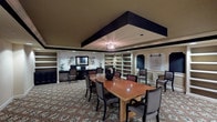 This single-floor multipurpose meeting and business center is designed to support both collaborative gatherings and individual productivity. The space is anchored by a large rectangular conference table surrounded by upholstered chairs, making it well-suited for group meetings or presentations. A distinctive tray ceiling with bold dark trim and a central chandelier-style light fixture adds architectural character and a sense of refined structure to the room.

Along one wall, a dedicated workstation area features dark-finished cabinetry, built-in desks equipped with a computer and printer, open shelving, and decorative mirrors and wall art — providing a functional business center setup within the same space. Opposite, built-in open shelving units frame framed chalkboard panels, offering flexible display and writing surfaces. Motivational wall lettering reading "Work Hard and Stay Humble" adds an inspirational touch to the environment.

The flooring throughout is covered in a richly patterned carpet with floral and botanical motifs, lending warmth and visual interest. Recessed lighting supplements the ambient illumination, ensuring the space is well-lit throughout. The overall design balances practicality with polished, hospitality-inspired aesthetics, creating a professional yet inviting atmosphere suitable for a variety of uses.