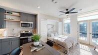 This thoughtfully designed single-floor studio maximizes every inch of its open layout through smart, built-in solutions. The main living area centers around a white Murphy bed system flanked by integrated cabinetry and drawers, allowing the space to transition seamlessly between sleeping and living configurations. Light hardwood flooring runs throughout, complementing the neutral white walls and contributing to a bright, cohesive aesthetic. A ceiling fan with a light fixture overhead aids in air circulation and adds a contemporary touch.

Large windows on the exterior wall flood the space with natural light and provide views of the surrounding neighborhood. A glass door opens directly onto a private balcony, further enhancing the sense of openness. The kitchen is fully equipped and efficiently arranged in an L-shape, featuring gray flat-front cabinetry extending to the ceiling, stainless steel appliances including a refrigerator, electric range, built-in microwave, and dishwasher, along with a white subway tile backsplash and open floating shelves for additional storage.

The bathroom maintains a consistent modern palette, featuring gray cabinetry with brushed nickel hardware, a white quartz countertop, an undermount sink, and a large mirror above the vanity. Dark tile flooring grounds the space with contrast.