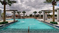 Located at 300 Bruce St in Conway, Arkansas, this expansive outdoor amenity space is designed to deliver a resort-style experience. The centerpiece is a large, vivid aqua swimming pool flanked by elegant concrete pergola columns from which graceful water jets arc into the pool, creating a lively and visually striking atmosphere. Tall palm trees are positioned throughout the deck, adding a tropical aesthetic that complements the open-sky setting.

Surrounding the pool, a generously sized concrete deck provides ample room for relaxation and socializing. Rows of wicker chaise lounges with plush gray cushions are arranged along the poolside, offering comfortable sunbathing areas. String lights stretch across portions of the space, lending a warm ambiance during evening hours.

Adjacent covered pavilions with brick and stone construction provide shaded retreat areas furnished with outdoor seating, ideal for gatherings. A covered veranda with white colonial-style columns and an ornate wrought iron balcony above is attached to the main building, extending the usable outdoor living area. Additional lounge groupings and a spa area are visible throughout the grounds. The overall design balances upscale architectural detailing with relaxed, resort-inspired comfort, making this outdoor space a standout community amenity.
