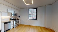This thoughtfully designed single-floor unit offers a clean, modern aesthetic throughout. Light hardwood flooring extends seamlessly across all living areas, creating a warm and cohesive foundation for the space. Black-framed windows frame views of the surrounding urban brick architecture, allowing natural light to filter in and connect the interior to its city setting.

The kitchen is well-appointed with stainless steel appliances, including a range, over-the-range microwave, and dishwasher. White cabinetry with black hardware reaches toward the ceiling, maximizing storage, while a classic white subway tile backsplash and granite countertops add refined detail. A geometric recessed ceiling light fixture adds a contemporary design touch overhead.

The bathroom is finished with large-format gray floor tiles and white subway tile wainscoting that lends a timeless, clean look. A wall-mounted vanity with a rich wood cabinet base, a mirrored medicine cabinet, and chrome fixtures complete the space. Accessibility grab bars are thoughtfully incorporated as well.

The remaining rooms are open and airy, each featuring the same consistent hardwood flooring and black-framed windows. The layout flows efficiently through a hallway connecting the rooms, with a bold black entry door providing a striking contrast to the bright white walls and trim throughout the unit.