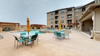 This outdoor amenity space is centered around a generously sized swimming pool with a classic rectangular design and a raised spa that flows water into the main pool via a spillover feature. The pool deck is finished in light-toned concrete, providing ample space for lounging and social gathering. Rows of teal chaise lounges line the poolside, while multiple dining tables with teal chairs and yellow market umbrellas are distributed throughout the patio for shaded seating.

A stone-clad fire pit is positioned on the open deck, serving as a focal point for outdoor gatherings. Toward one end of the courtyard, a wood-beamed pergola shelters an outdoor kitchen area equipped with granite countertops and stone columns, providing a covered space for cooking and entertaining. Additional pergola structures are placed along the perimeter, adding architectural definition to the space.

The courtyard is enclosed by a multi-story brick and stucco building with private balconies overlooking the amenity area. Landscaped planters with ornamental grasses accent the building edges. The overall design creates a cohesive, resort-inspired outdoor environment well-suited for relaxation and social activity.