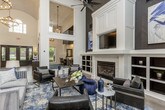 This striking single-floor space makes an immediate impression with its grand, double-height entrance foyer, where dramatic black-framed French doors and ornate iron-accented entry doors usher guests inside beneath a statement chandelier. Soaring ceilings flood the open interior with natural light through an abundance of large, multi-pane windows that frame views of lush greenery outdoors.

Throughout the space, wide-plank hardwood flooring in a warm, light-toned finish runs continuously, unifying the various zones seamlessly. The main lounge area is anchored by a bold dark herringbone feature wall that rises dramatically to the ceiling, providing a striking backdrop for curated artwork and paired table lamps. Navy blue velvet seating arrangements are thoughtfully grouped around a round stone-topped cocktail table, creating an inviting and sophisticated atmosphere.

A secondary lounge area near a white fireplace surround with built-in shelving offers additional seating with leather chairs and a layered area rug. A long hallway connects the lounge spaces to a well-appointed kitchen and dining area featuring pendant lighting, dark cabinetry, and bar-height seating. Structural columns with dark accents punctuate the open floor plan, adding architectural definition. The overall design blends modern elegance with welcoming warmth throughout.