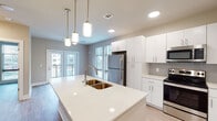This 1-bedroom, 1-bathroom residence at The Oliver in North Little Rock, Arkansas, offers a thoughtfully designed, single-floor layout with contemporary finishes throughout. The open-concept kitchen serves as the heart of the home, anchored by a generous quartz-topped island with a double basin sink and elegant pendant lighting. Crisp white cabinetry extends floor to ceiling, complemented by a full suite of stainless steel appliances including a refrigerator, range, and built-in microwave. A tile backsplash adds a refined touch, while light wood-style flooring carries seamlessly throughout the space. The living area extends naturally from the kitchen, bathed in natural light from large windows and French doors that open to an outdoor balcony, creating an airy and inviting atmosphere. The bedroom is generously proportioned, featuring a ceiling fan and a wide bank of windows that frame outdoor views. The bathroom is cleanly appointed with a long white vanity offering ample storage, light wood flooring, and access to a dedicated walk-in closet, providing excellent organization and comfort. Neutral warm tones, recessed lighting, and clean architectural lines give the entire residence a polished, modern feel.