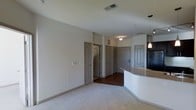 This single-floor residence offers a clean, open-concept layout designed for comfortable everyday living. The main living area is finished with plush carpeting in a neutral tone, creating a warm and inviting atmosphere, while the adjacent hallway and entry zone transition to rich, dark hardwood flooring that adds a contrasting elegance to the space.

A standout architectural feature is the curved kitchen peninsula, finished with a light stone-toned countertop and accompanied by a stylish pendant light overhead. The curved design adds a modern, flowing quality to the kitchen boundary, seamlessly connecting the cooking area to the living space.

The unit features multiple interior doors with arched panel detailing in a soft gray finish, lending a subtle traditional character to the otherwise contemporary design. These doors lead to additional rooms and closets, suggesting a thoughtfully segmented floor plan that balances open living with private spaces.

The ceilings are smooth and well-proportioned, with recessed ventilation and flush lighting fixtures that keep the aesthetic uncluttered. Neutral wall tones throughout the space provide a versatile backdrop, allowing for a wide range of interior styling. Overall, the space presents a well-balanced blend of modern convenience and classic design touches.