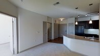 This single-floor residence offers a clean, open-concept layout designed for comfortable everyday living. The main living area is finished with plush carpeting in a neutral tone, creating a warm and inviting atmosphere, while the adjacent hallway and entry zone transition to rich, dark hardwood flooring that adds a contrasting elegance to the space.

A standout architectural feature is the curved kitchen peninsula, finished with a light stone-toned countertop and accompanied by a stylish pendant light overhead. The curved design adds a modern, flowing quality to the kitchen boundary, seamlessly connecting the cooking area to the living space.

The unit features multiple interior doors with arched panel detailing in a soft gray finish, lending a subtle traditional character to the otherwise contemporary design. These doors lead to additional rooms and closets, suggesting a thoughtfully segmented floor plan that balances open living with private spaces.

The ceilings are smooth and well-proportioned, with recessed ventilation and flush lighting fixtures that keep the aesthetic uncluttered. Neutral wall tones throughout the space provide a versatile backdrop, allowing for a wide range of interior styling. Overall, the space presents a well-balanced blend of modern convenience and classic design touches.