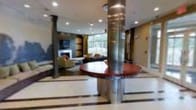 This elegant single-floor space presents a refined and welcoming atmosphere, characterized by a blend of modern and classic design elements. At the heart of the space stands a prominent circular reception desk with a rich, dark wood finish, anchored by a sleek polished metal column that extends to the ceiling — a clear architectural focal point that commands attention upon entry.

The flooring features a light-toned tile with contrasting inlay details, adding a sense of structure and formality to the layout. Additional decorative columns with a dark finish frame the space, complementing the warm, neutral tones of the surrounding walls.

A comfortable lounge area is situated toward the rear of the space, furnished with upholstered seating arranged around a classic white fireplace, creating an inviting retreat for guests. Built-in shelving and wall paneling add depth and a sense of permanence to this area.

Floor-to-ceiling windows along the back wall flood the lounge with natural light and offer views of lush greenery outside, seamlessly connecting the interior to the natural environment. Recessed ceiling lighting throughout ensures the space remains well-lit and polished at all times. Overall, the space conveys a professional yet comfortable ambiance suited to a hospitality or corporate environment.