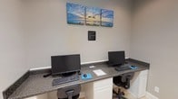 This business center at 14510 Lemoyne Blvd in Biloxi, Mississippi, offers a compact yet well-equipped space designed for productivity. The room features light gray walls, clean white baseboards, and light-toned tile flooring, creating a bright and professional atmosphere. Recessed ceiling lights provide consistent, focused illumination throughout the space.

The centerpiece of the room is a built-in granite-topped workstation along one wall, accommodating two desktop computer setups complete with monitors, keyboards, and ergonomic task chairs. White cabinetry beneath the countertop adds practical storage while maintaining a polished appearance. A posted Business Center Rules sign keeps the space organized and user-friendly.

Adorning the wall above the workstations is a striking triptych canvas print depicting a coastal sunset scene — a fitting nod to the Gulf Coast location that adds a touch of local character to the room.

A freestanding bookshelf in the corner serves as a free mini library, stocked with a varied collection of books available for guests to enjoy. The room is accessed through a classic six-panel white interior door, reinforcing the space's clean, traditional aesthetic.

Overall, this business center provides a quiet, functional environment well-suited for work, browsing, or leisure reading.