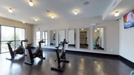 This well-appointed fitness studio offers a clean, contemporary environment designed to support a variety of workout routines. The space is anchored by rich, dark hardwood flooring that runs throughout, providing both durability and a polished aesthetic. A row of framed wall mirrors lines one side of the room, maximizing the sense of space and allowing exercisers to monitor their form during workouts. The white brick-textured accent wall adds a subtle industrial character that complements the modern design. Several stationary cycling bikes are positioned across the floor, creating a dedicated spin area with ample room between each station. Flush-mount ceiling lights and wall-mounted fixtures provide bright, even illumination throughout the studio. Toward the rear of the space, an adjoining room serves as a dedicated storage and equipment zone, visible through the mirrors and outfitted with shelving units and a colorful selection of fitness accessories, including resistance bands and weighted balls. A wall-mounted speaker system supports an energizing workout atmosphere. The overall ambiance is clean, functional, and motivating, making this fitness studio a well-equipped destination for both individual training and group exercise sessions.