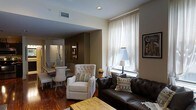 This single-floor space offers a thoughtfully arranged open-concept layout that seamlessly connects the living and dining areas. The living room is anchored by a rich, dark leather sofa accented with patterned throw pillows, complemented by a tufted white accent chair that adds a touch of classic elegance. A rustic wooden coffee table grounds the seating arrangement, adding warmth and texture to the space.

Tall windows dressed in sheer white curtains allow natural light to filter softly into the room, highlighting the rich hardwood flooring that runs throughout. The neutral wall tones in warm beige and white create a cohesive backdrop that enhances the inviting atmosphere.

Adjacent to the living area, the dining space features a light-toned table with coordinating chairs, making it well-suited for everyday dining. A decorative wall hanging and framed artwork, including what appears to be a vintage city-themed print, add character and a sense of place to the interior.

Recessed lighting supplements the natural light, ensuring the space remains bright and functional throughout the day. Overall, the design strikes a balance between comfort and style, with carefully chosen furnishings that contribute to a cozy yet refined atmosphere.