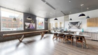 Located at 2101 Market St in Denver's vibrant RiNo neighborhood, this single-floor amenity space is thoughtfully designed to serve as a social and recreational hub. The open-plan layout seamlessly connects several distinct zones, each finished with wide-plank hardwood-style flooring, a bold charcoal ceiling, and an eclectic mix of industrial and mid-century modern furnishings.

The game and entertainment zone anchors one side of the space, featuring a full-length shuffleboard table, large wall-mounted flat-screen TVs, and striking chevron-patterned dark wall panels. Pendant lighting in matte black and brushed gold casts warm, focused light throughout. Oversized windows along the exterior walls flood the area with natural light while offering direct views of the surrounding cityscape and Denver's colorful street art.

A communal dining area sits at the center, furnished with a large oval table and mid-century style chairs, ideal for group gatherings. Adjacent to it is a well-appointed kitchenette with white subway tile backsplash, wood cabinetry, and counter seating. A cozy lounge nook with curved walls and plush gray accent chairs provides a quieter retreat, accented by curated artwork and sculptural décor. A glazed partition wall connects the main lounge to a fully equipped fitness center, keeping the space bright and visually expansive.