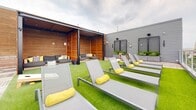 Located at 1229 Chapel St in New Haven, CT, this rooftop terrace at Novella offers a well-appointed outdoor retreat above the city. The space is thoughtfully designed, blending artificial turf, wood decking, and contemporary furnishings to create a versatile environment suited for both relaxation and social gatherings.

A row of sleek gray lounge chairs with bright yellow accent pillows lines the turf area, providing ample space for sunbathing while overlooking the New Haven skyline. A wood-clad cabana structure anchors one end of the terrace, offering a shaded lounge area fitted with modular sectional seating and decorative pillows—ideal for intimate gatherings or casual lounging.

On the opposite end, a dedicated outdoor kitchen and bar area features a built-in grill, wood-front bar counter with seating, and a wall-mounted television, making it a natural hub for entertaining. A cornhole set adds a recreational touch to the open turf zone. Planter boxes with greenery soften the perimeter walls, and cable railing along the terrace edges preserves unobstructed panoramic views of the surrounding New Haven cityscape. The overall ambiance is modern and inviting, combining urban energy with rooftop comfort.
