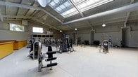 This fitness facility is designed to accommodate a wide range of training needs within a single, expansive floor. The gym's most striking architectural feature is its large skylight system running across the ceiling, which floods the workout floor with generous natural light throughout the day, complemented by pendant lighting fixtures for consistent illumination during all hours.

The ceiling is notably high, with an industrial-style design that incorporates exposed ductwork and structural beams, contributing to an open and airy atmosphere. The neutral-toned walls and light concrete flooring create a clean, functional aesthetic throughout the space.

The workout floor is generously proportioned, housing an extensive array of strength training machines, including cable systems, resistance machines, and weight benches, arranged to allow comfortable movement between stations. A dedicated free weights area features a full rack of dumbbells along one wall, while additional stations are distributed across the floor to maximize usability.

Windows along the upper walls further enhance natural ventilation and brightness, reinforcing the spacious feel of the facility. The overall layout is thoughtfully organized, offering clear pathways between equipment zones. This facility is well-suited for both individual training sessions and group fitness use, combining practical design with a professional-grade equipment setup.