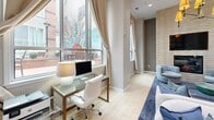 Located at 401 Granby St in the heart of downtown Norfolk, Virginia, this single-floor space offers a thoughtfully designed open-plan layout that seamlessly integrates a living area and a fully equipped kitchen. The living room is anchored by a striking floor-to-ceiling stacked stone accent wall housing a fireplace and wall-mounted television, creating a bold focal point. A contemporary chandelier with amber shades hangs overhead, complementing the recessed lighting that illuminates the space throughout. Large, floor-to-ceiling windows dressed with flowing neutral-toned drapes flood the interior with natural light and frame views of the surrounding urban streetscape. Light-toned tile flooring runs throughout, enhancing the bright and airy atmosphere. A dedicated workspace with a glass-top desk is positioned near the windows, taking advantage of the natural light. The kitchen features warm wood cabinetry, striking blue-veined granite countertops extending along a generous bar-height island, a glass mosaic tile backsplash, and stainless steel appliances. Contemporary artwork adorns the walls, adding vibrant pops of color. The overall ambiance is modern and polished, balancing comfort and sophistication in a prime downtown Norfolk location.